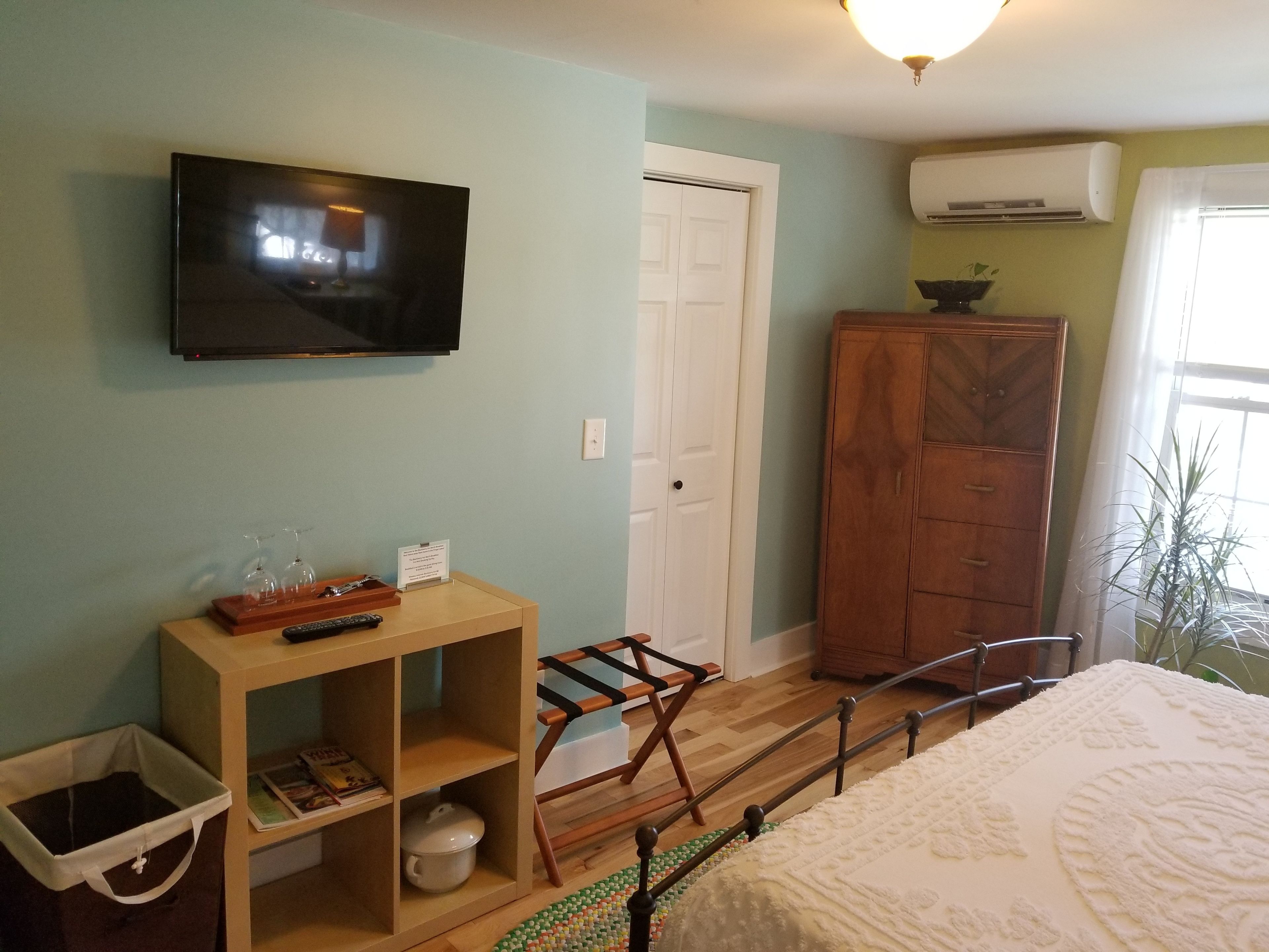 A bedroom with a bed, a TV mounted on the wall, a wooden cabinet, a small table with a remote control, and a window with a plant.