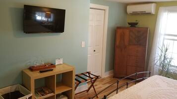 A bedroom with a bed, a TV mounted on the wall, a wooden cabinet, a small table with a remote control, and a window with a plant.