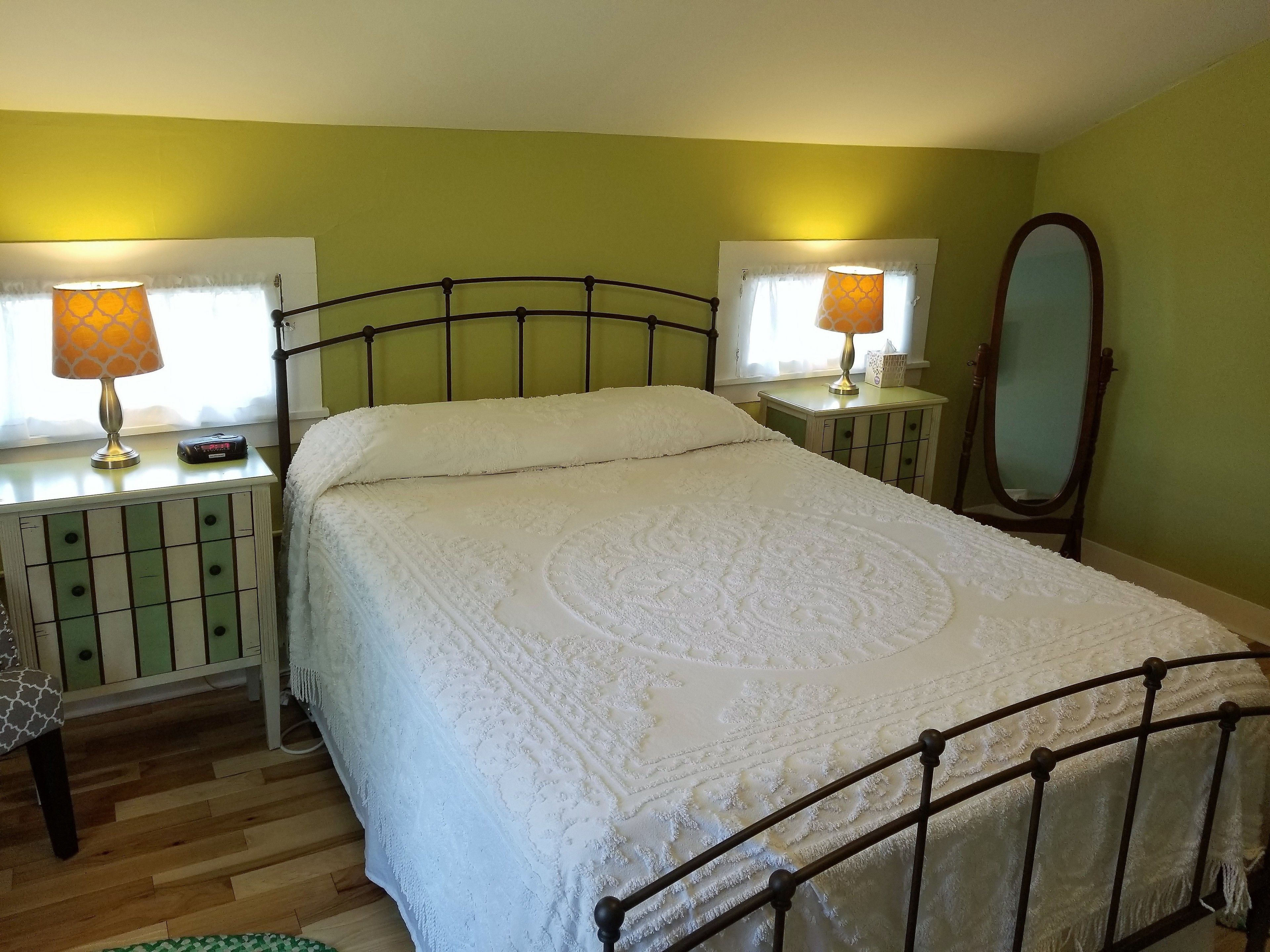 A neatly made bed with a white duvet, two bedside lamps, a bedside table with a lamp, a mirror, and a green wall.