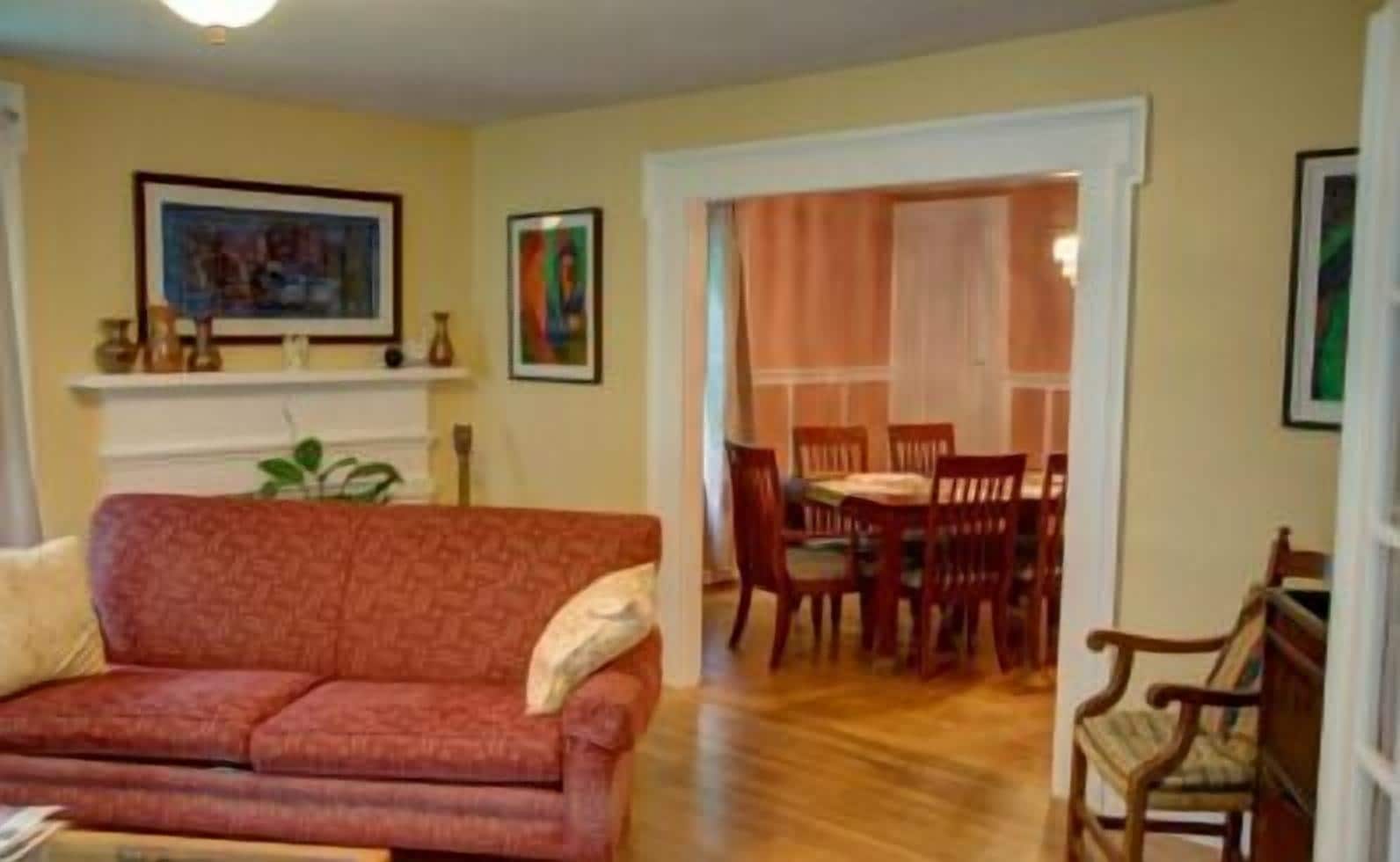 Ein Wohnzimmer mit rotem Sofa, einem Esstisch mit Stühlen und einem Kamin, über dem sich gerahmte Kunstwerke befinden.
