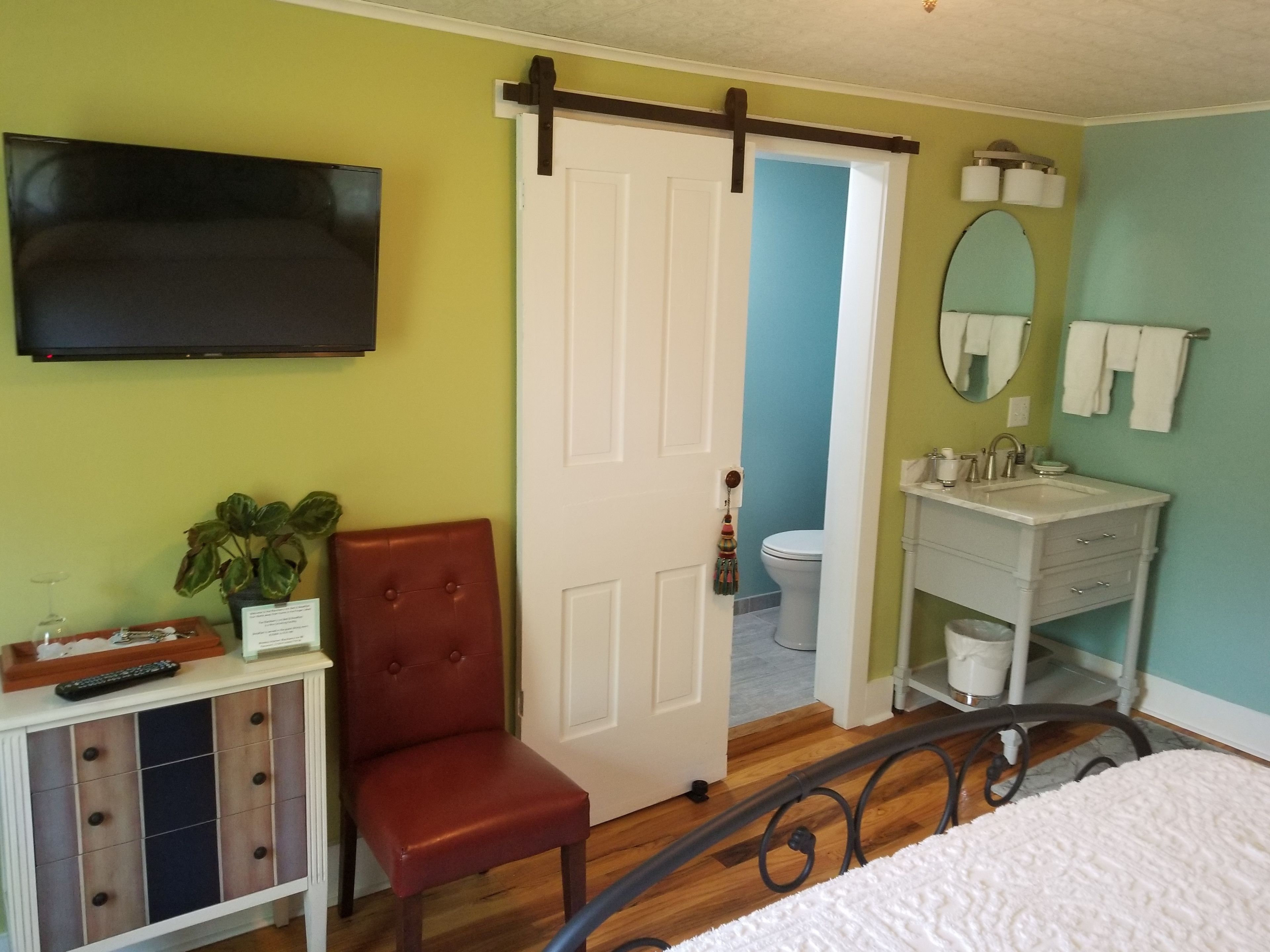Ein Schlafzimmer mit Bett, einem roten Sessel, einem Fernseher, einem Schminktisch und einem Badezimmer mit Toilette und Waschbecken. Eine Tür führt in einen anderen Raum.