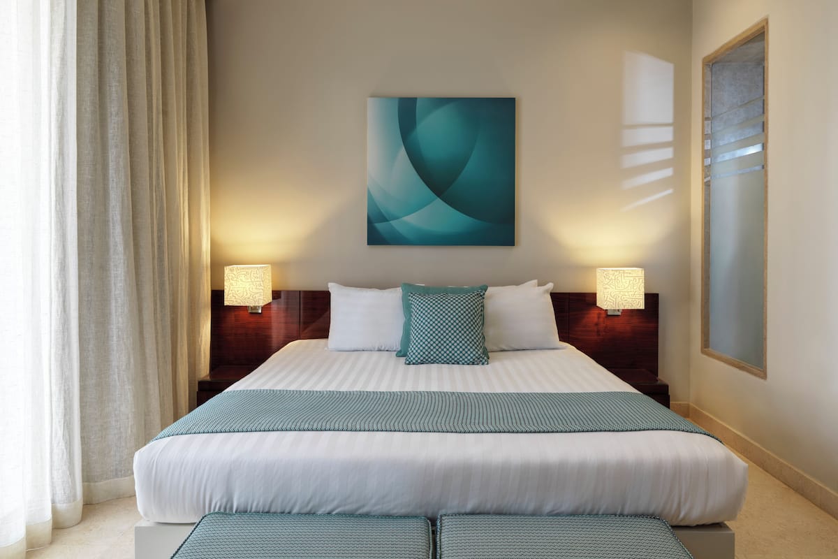 A hotel room with a large bed, two bedside lamps, a wall-mounted abstract painting, and a window with curtains.