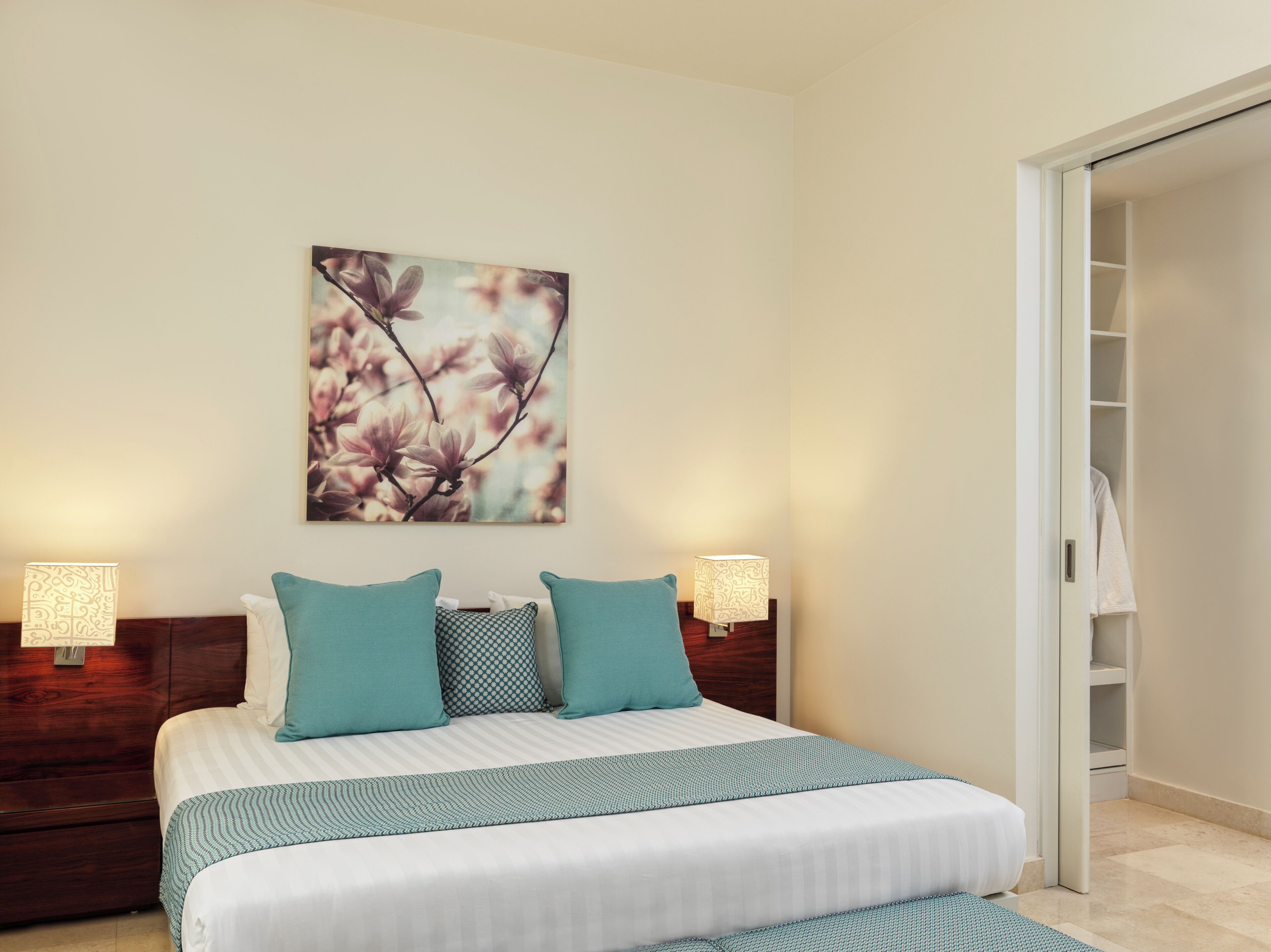 A neatly arranged bedroom with a bed, two bedside lamps, a wall-mounted artwork, and a wardrobe.