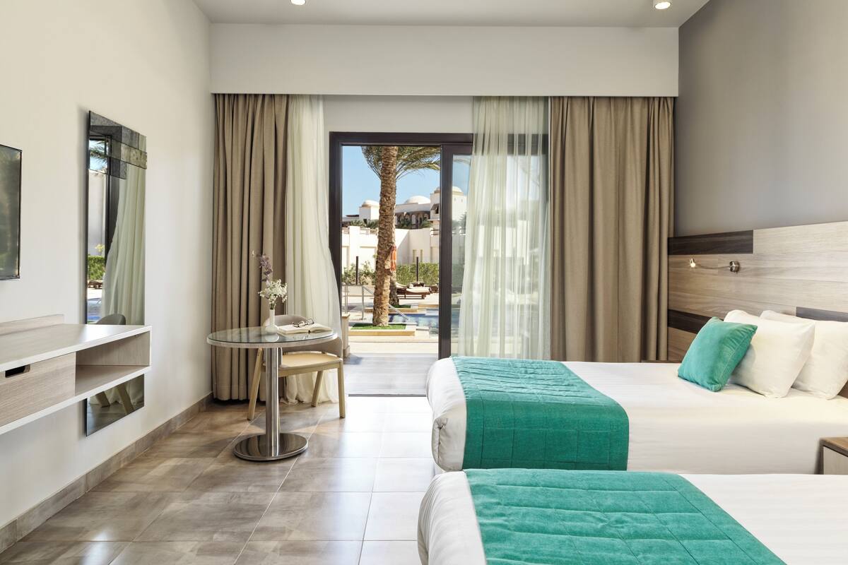 A hotel room with a bed, a desk, a chair, and a view of an outdoor pool and palm trees.
