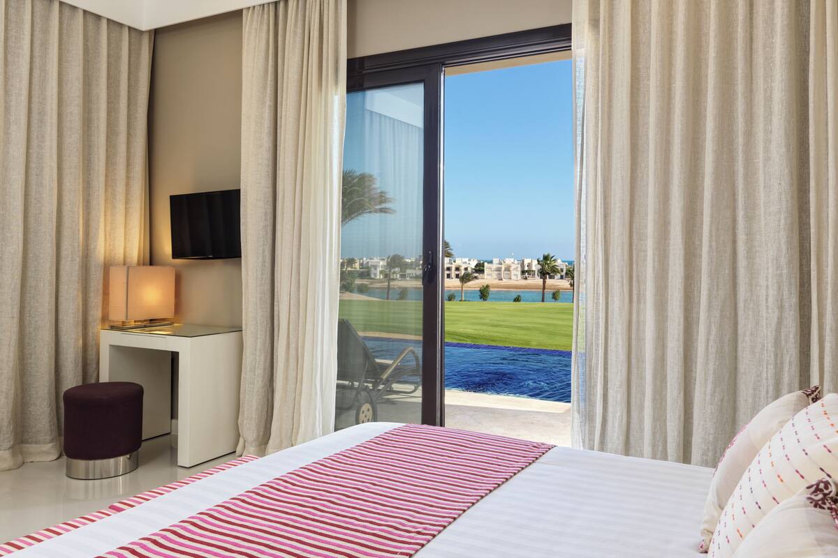 A hotel room with a bed, a desk with a lamp, a TV, and a view of the sea and buildings.