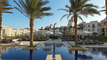 A pool area with lounge chairs and umbrellas, surrounded by palm trees and a building with multiple balconies.
