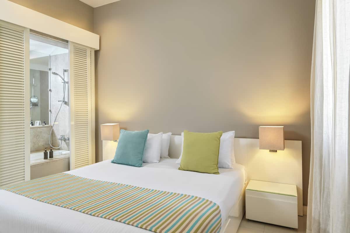 A hotel room with a large bed, two bedside tables, a window with blinds, and a bathroom with a shower and mirror.