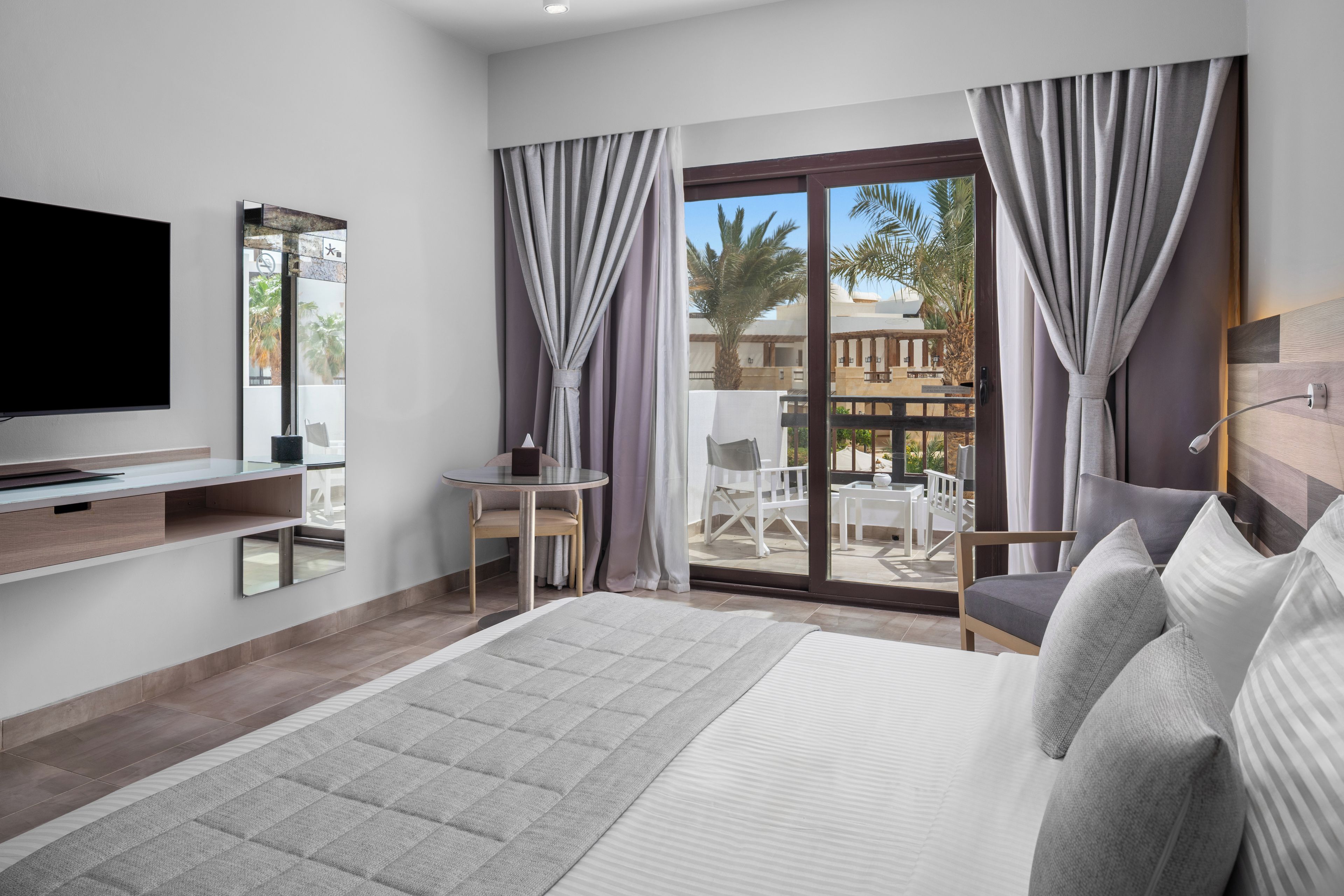 A modern hotel room with a large bed, a flat-screen TV, a small table, and a balcony with a view of palm trees.