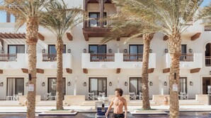 A man walking by a pool with palm trees and a building with balconies. - Ancient Sands Golf Resort and Residences (El Gouna)
