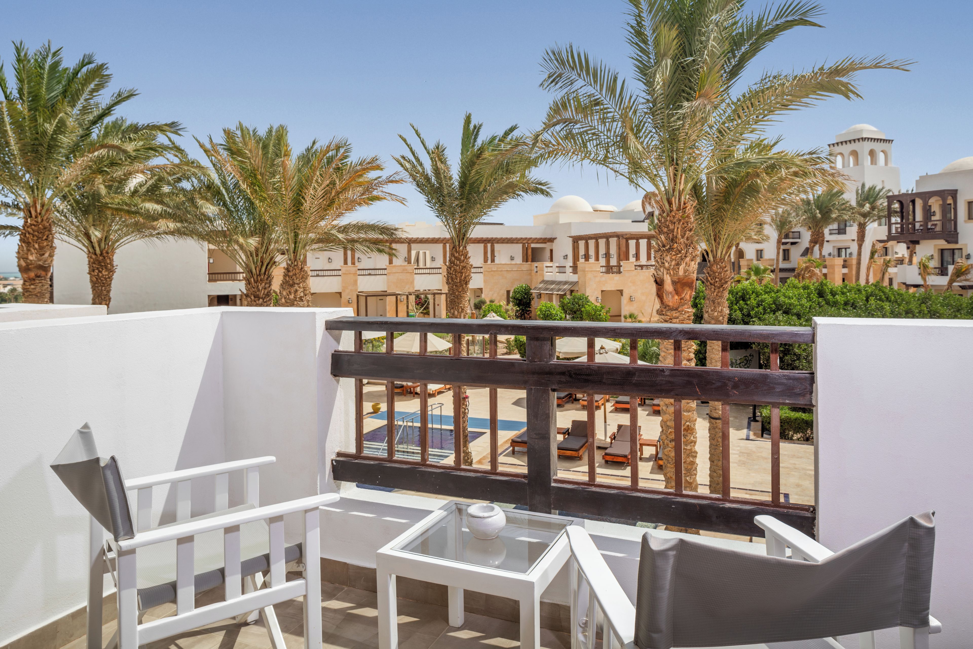 A balcony with white furniture, palm trees, and a view of a pool and buildings.