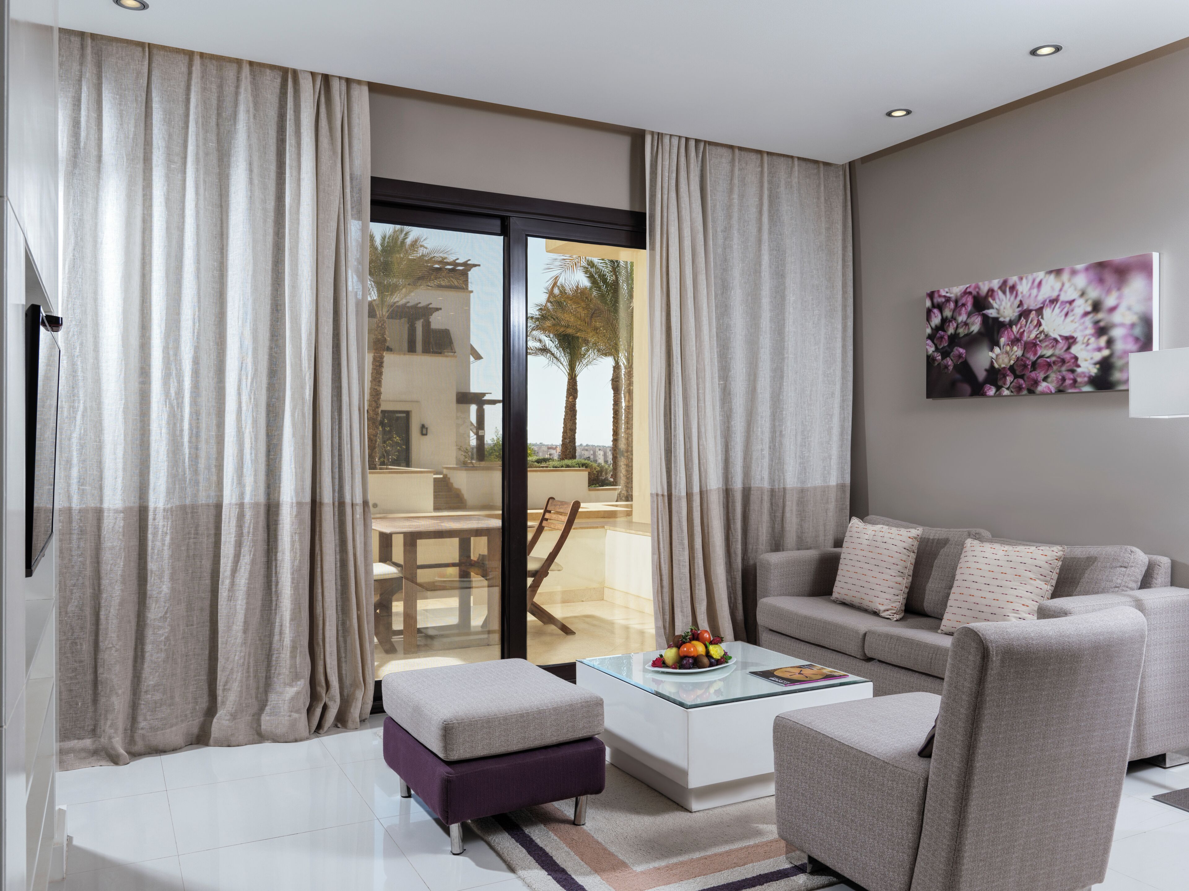 A modern living room with a sofa, ottoman, and coffee table. A glass door leads to an outdoor area with a patio and palm trees.