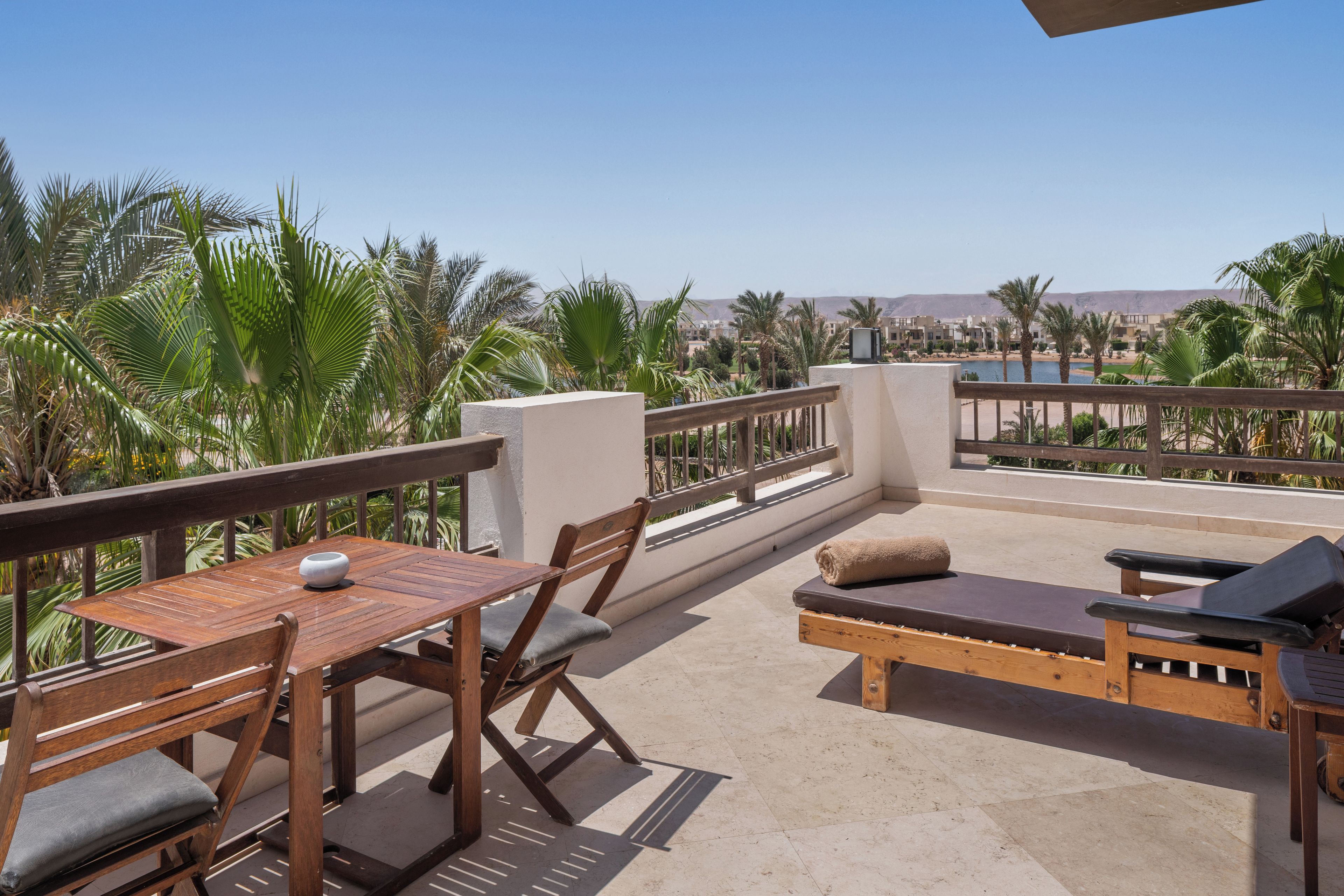 Una terraza con una mesa y sillas de madera, un sofá con cojines, y vistas a palmeras y edificios.
