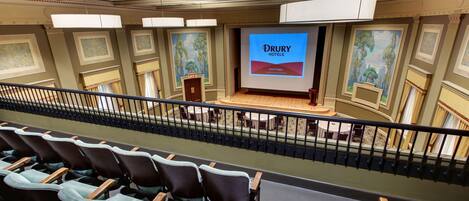 A Drury Hotel conference room with a large screen displaying the hotel's name, rows of chairs, and a balcony with railings.