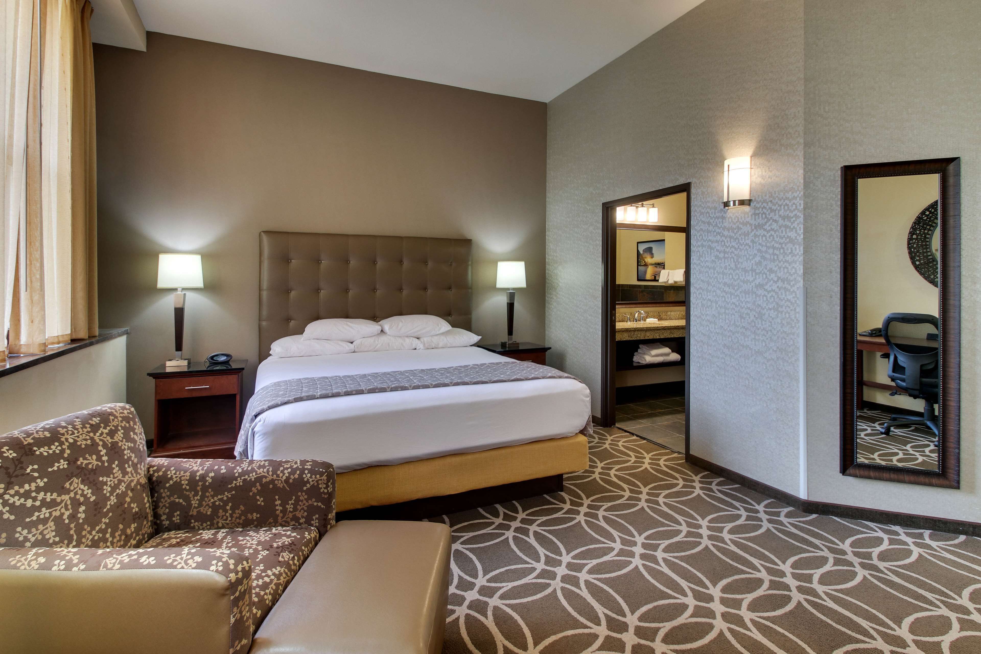 A hotel room with a large bed, a chair, a desk, a mirror, and a bathroom visible through an open door.