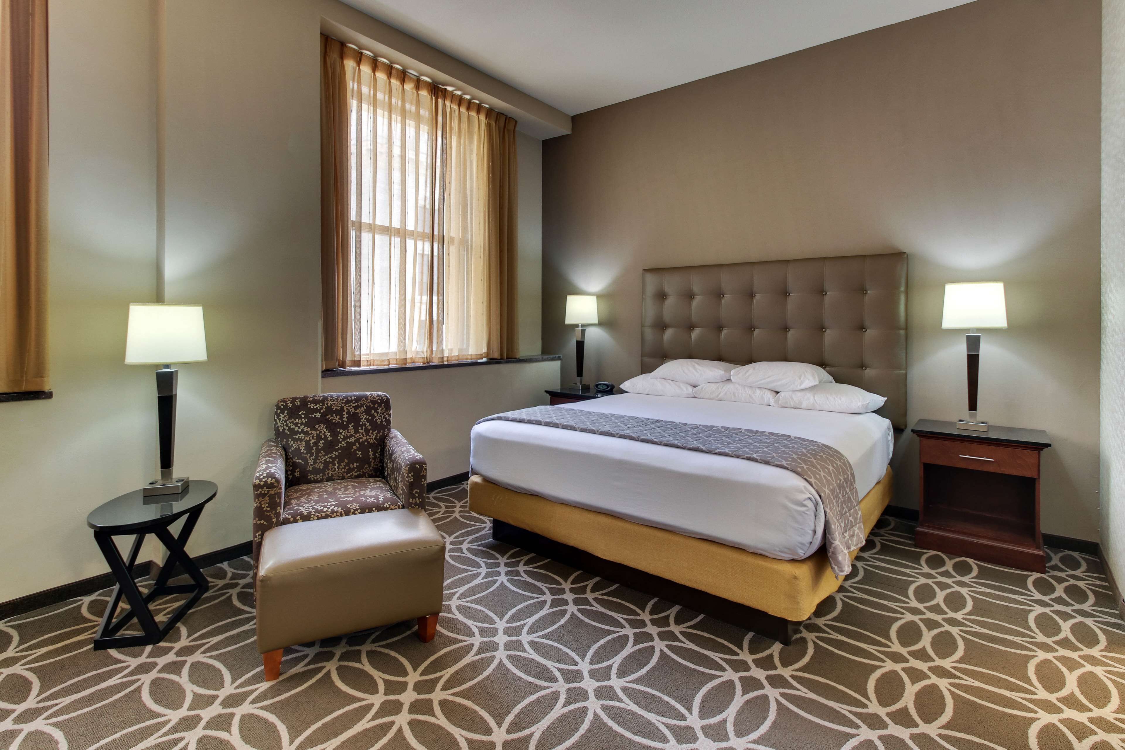 A hotel room with a bed, a chair, two nightstands with lamps, a window with curtains, and a patterned carpet.