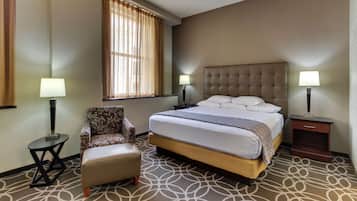 A hotel room with a bed, a chair, two nightstands with lamps, a window with curtains, and a patterned carpet.