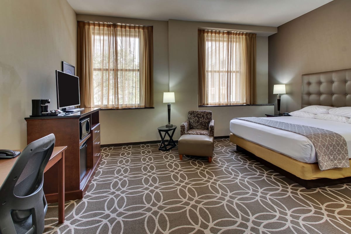 A hotel room with a bed, a desk with a computer, a chair, and two windows with curtains.