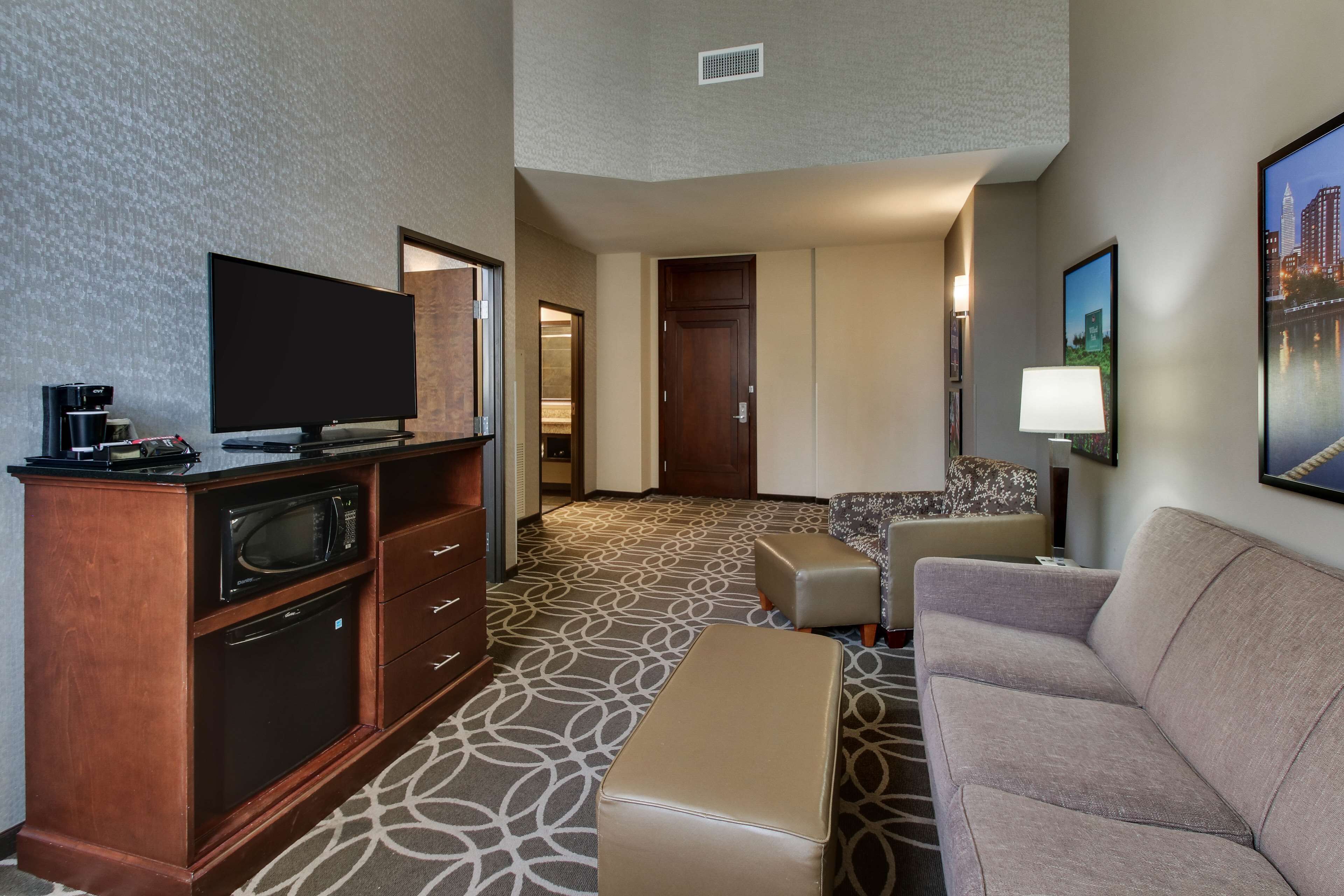A hotel room with a flat-screen TV, a minibar, a sofa, an ottoman, and a patterned carpet.