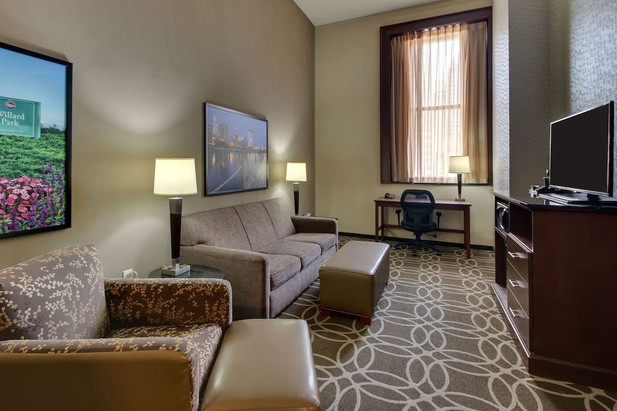 A hotel room with a sofa, armchair, ottoman, desk, television, and a large window with curtains.