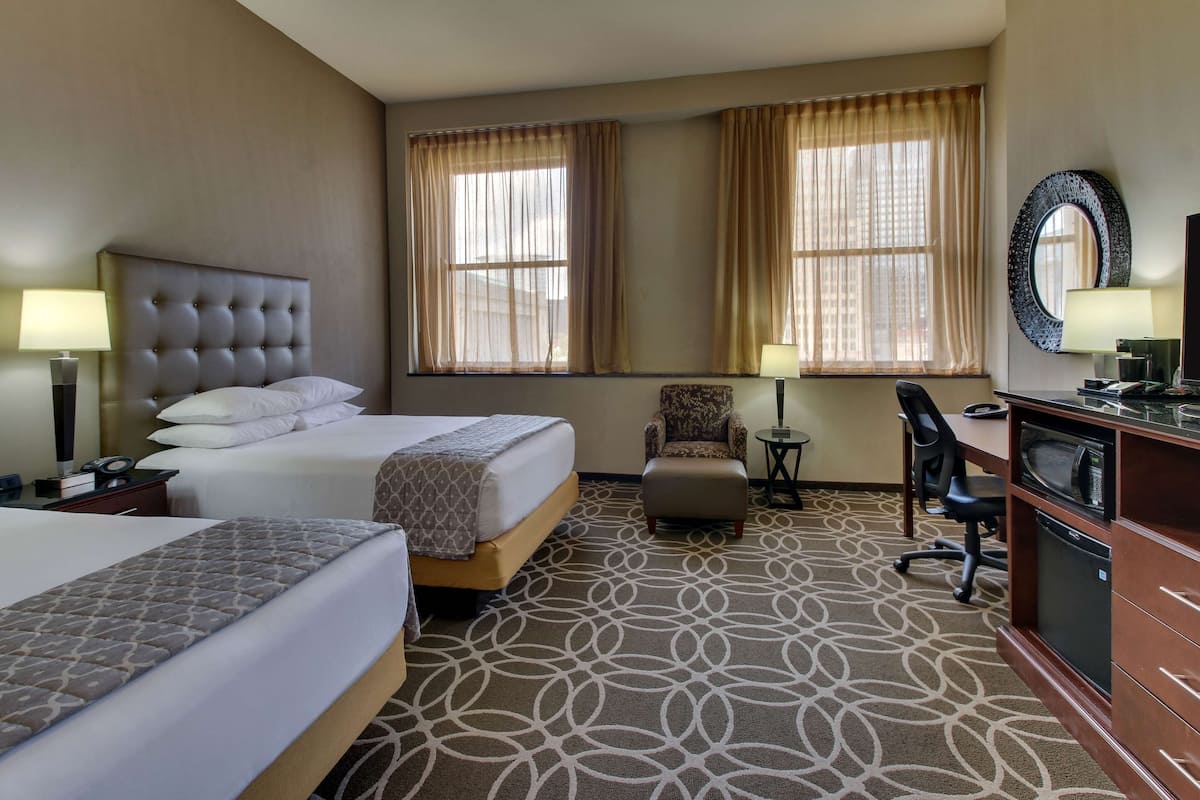 A hotel room with two beds, a desk, a chair, a TV, and a large window with curtains.