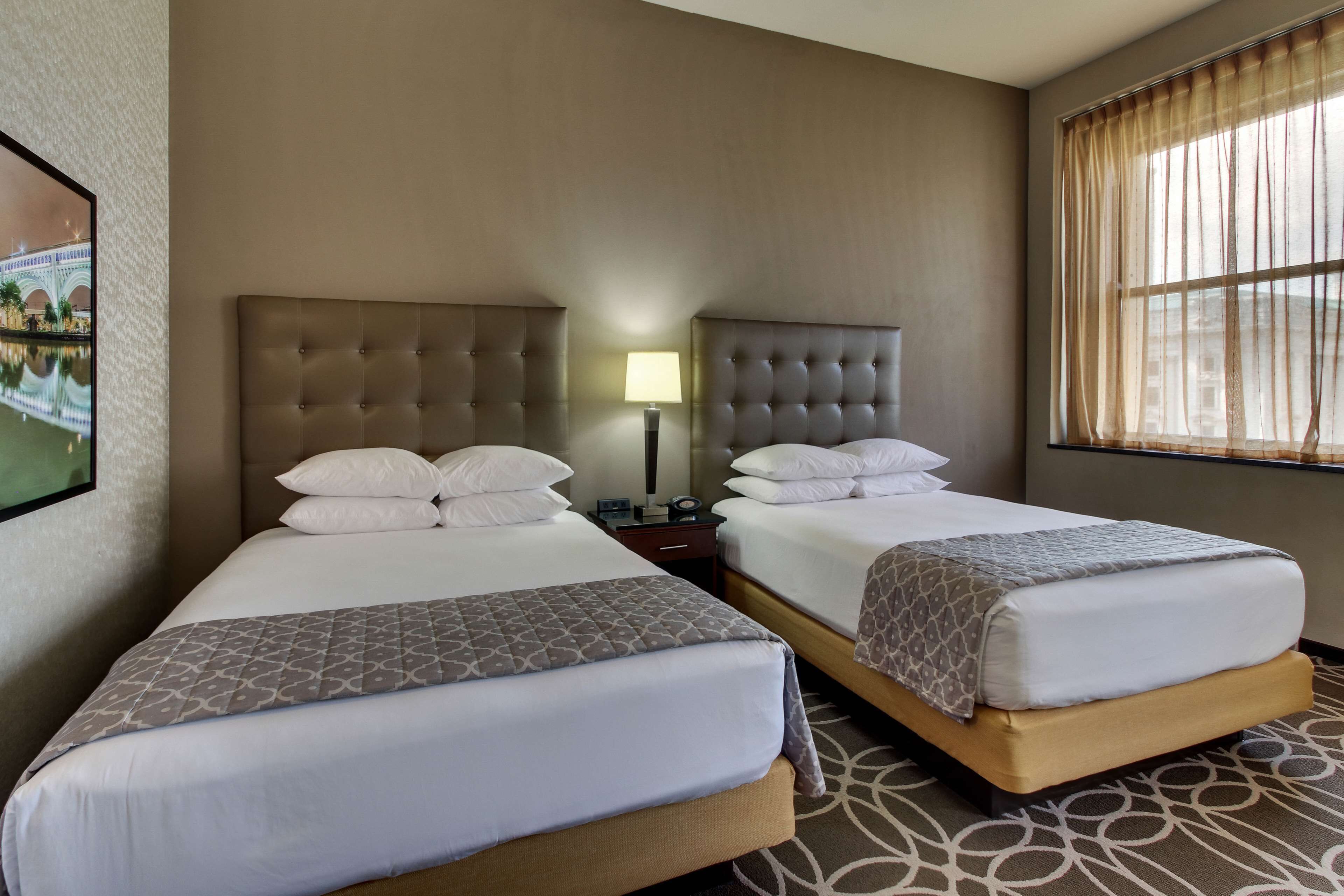 Habitación de hotel con dos camas, una mesita de noche, una lámpara y un ventanal con cortinas.