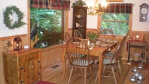 A wooden dining room with a large table set for six, surrounded by chairs. A chandelier hangs from the ceiling, and a clock is visible on the wall.