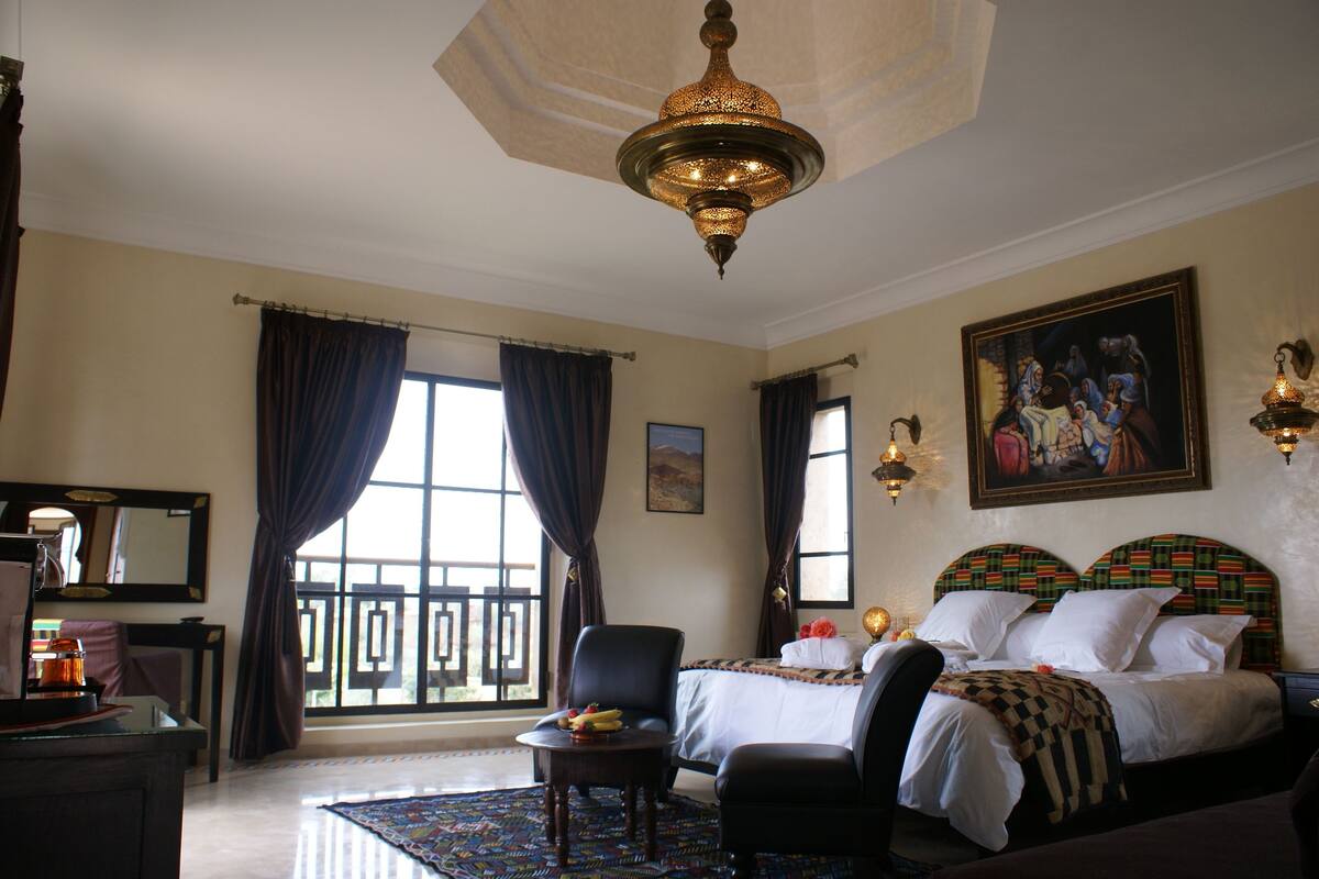 A hotel room with two beds, a desk, a chair, a large window with curtains, a chandelier, and artwork on the wall.