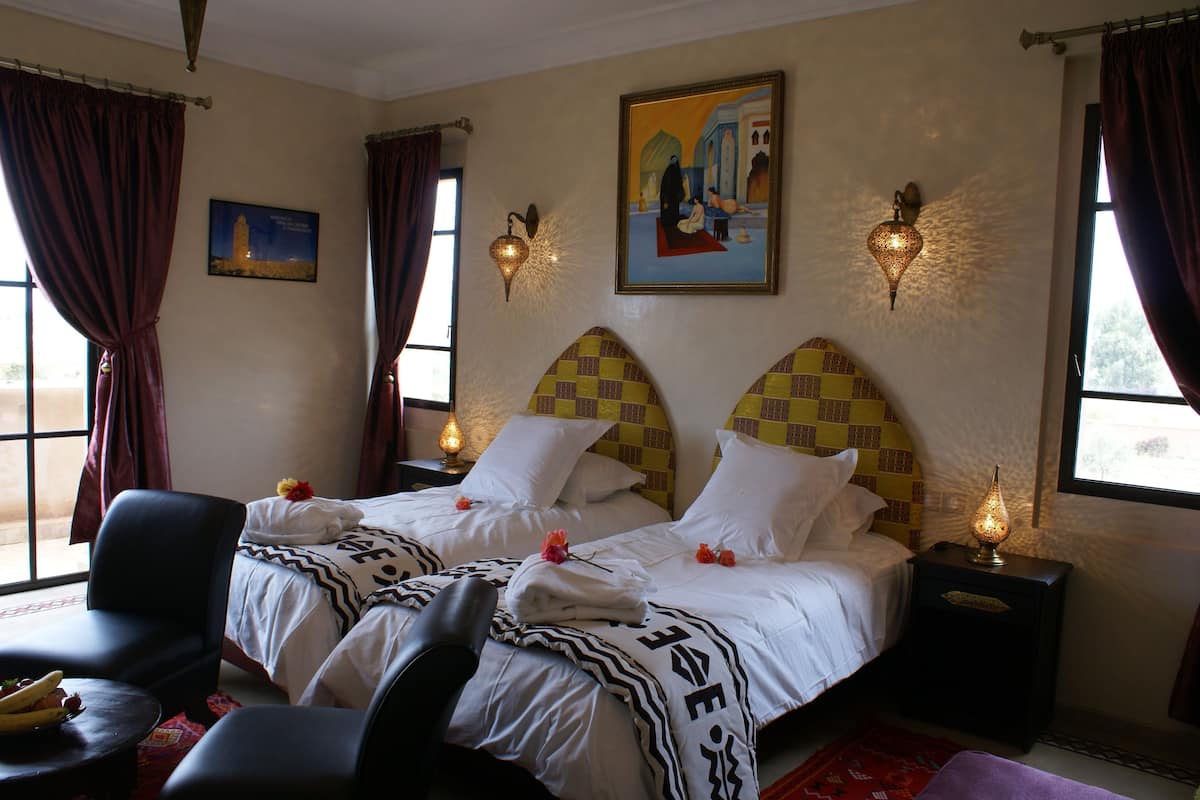 A hotel room with two beds, a chair, a nightstand with a lamp, and a painting on the wall.