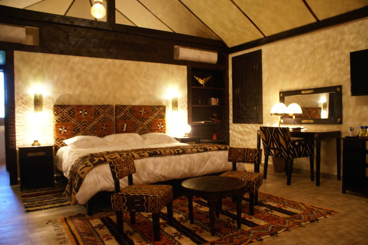 A bedroom with a large bed, a desk, and a chair. There is a rug on the floor and a lamp on the desk.