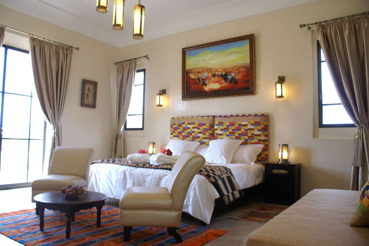A hotel room with a large bed, two bedside tables, a chair, a small table with fruit, and a painting on the wall.