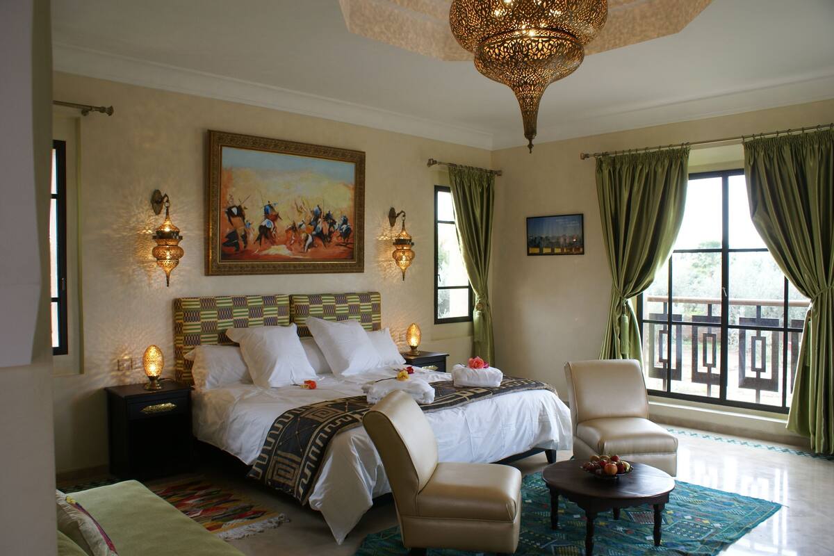 A hotel room with a large bed, two bedside tables with lamps, a chair, a small table with fruit, and a balcony view.