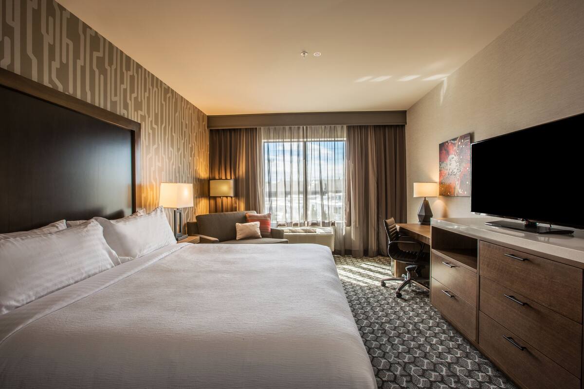 A hotel room with a large bed, a desk with a computer, a TV, and a window with curtains.