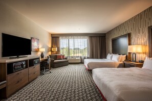 Een hotelkamer met een flatscreen-televisie, twee bedden, een bureau met lamp, een bank en een groot raam met gordijnen.