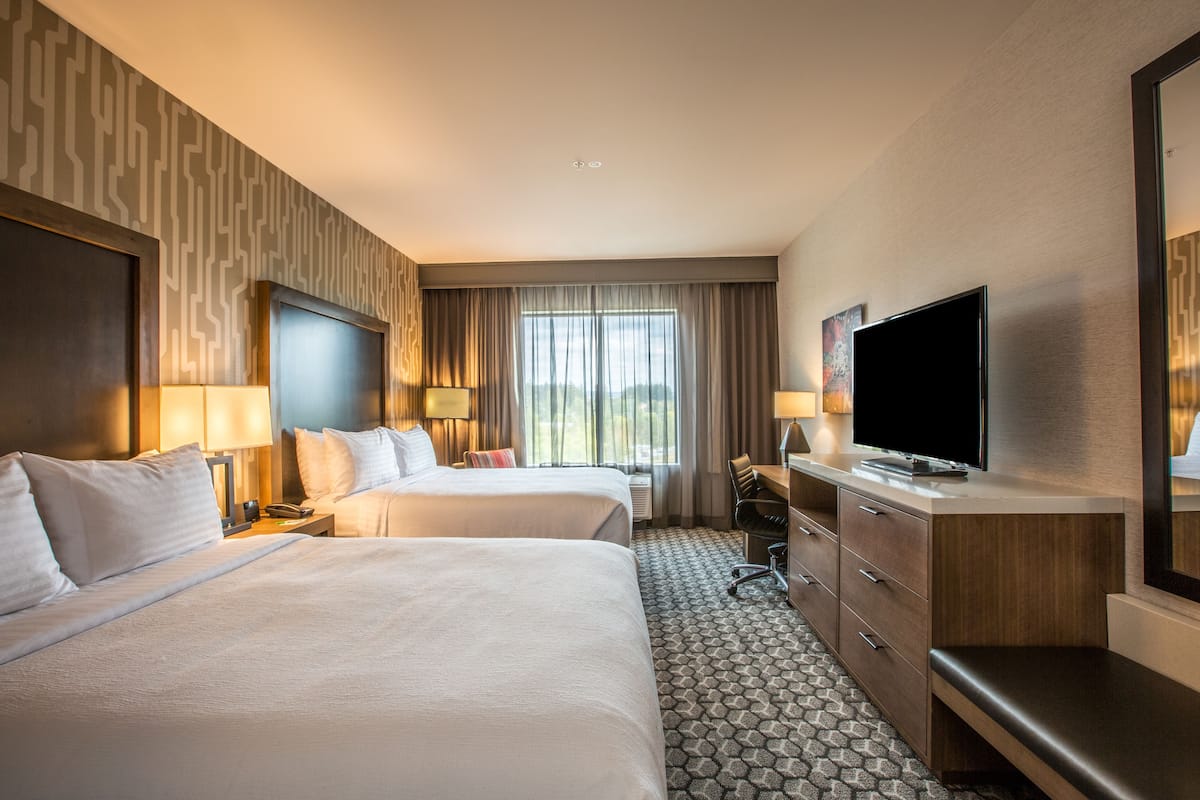 A hotel room with two beds, a desk, a television, and a large window with curtains.