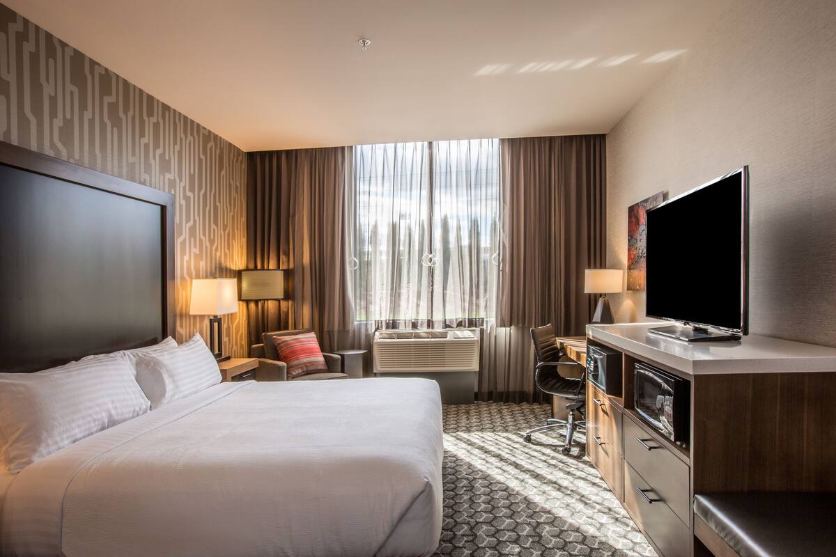 A hotel room with a large bed, a flat-screen TV, a desk with a chair, a window with curtains, and a wall-mounted air conditioning unit.