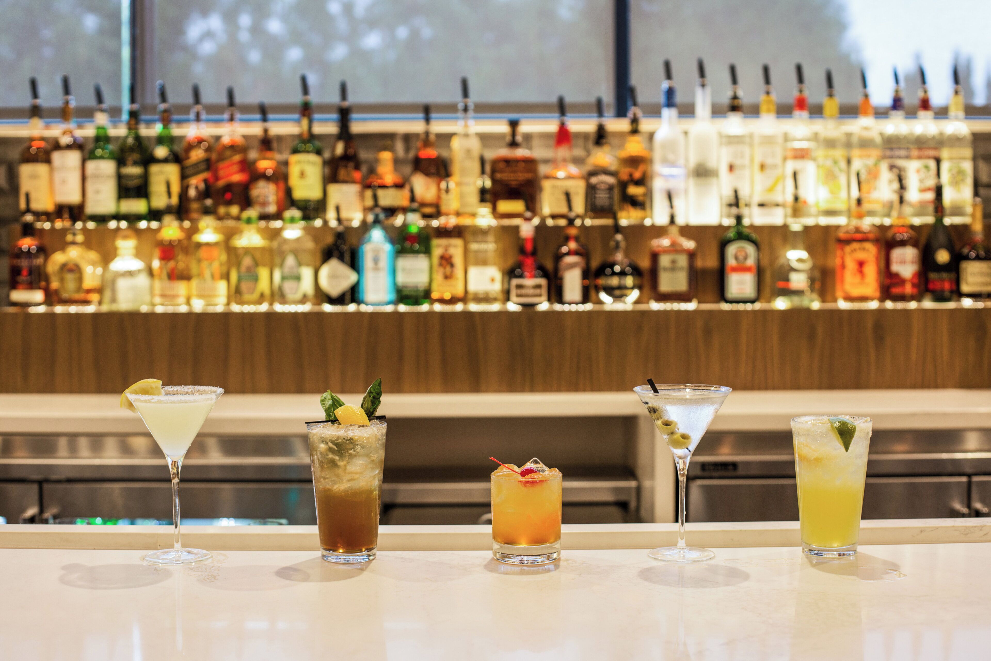 A bar with a variety of liquor bottles in the background and four different cocktails in the foreground.