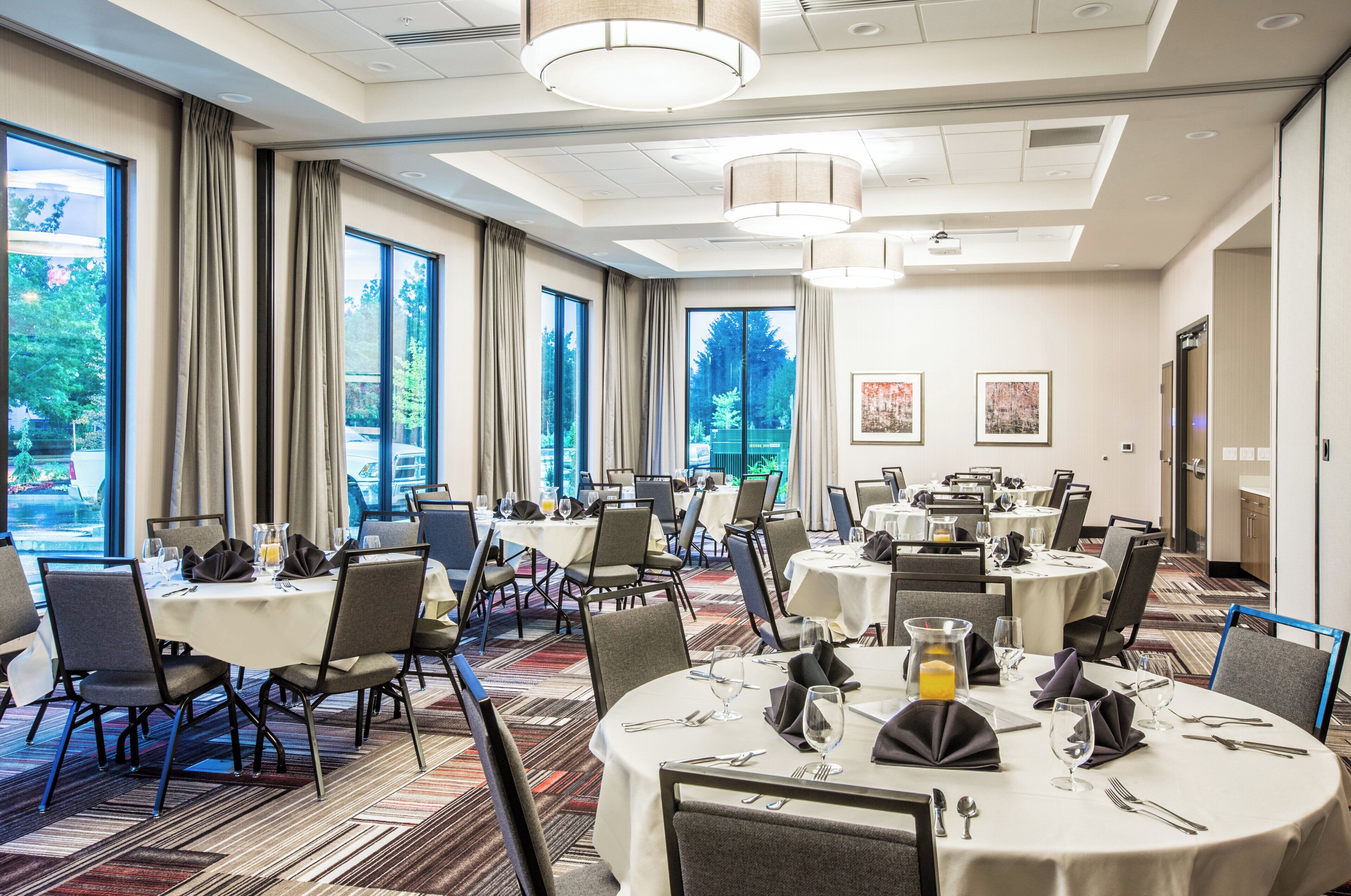 Un comedor de hotel con mesas redondas, sillas y grandes ventanales que ofrecen vistas al exterior.
