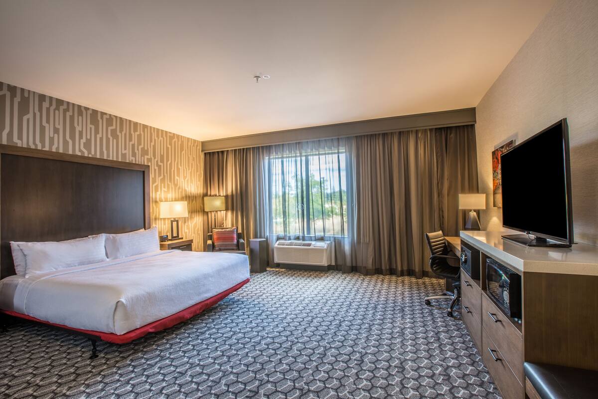 A hotel room with a large bed, a desk with a flat-screen TV, a desk lamp, a chair, and a window with curtains.