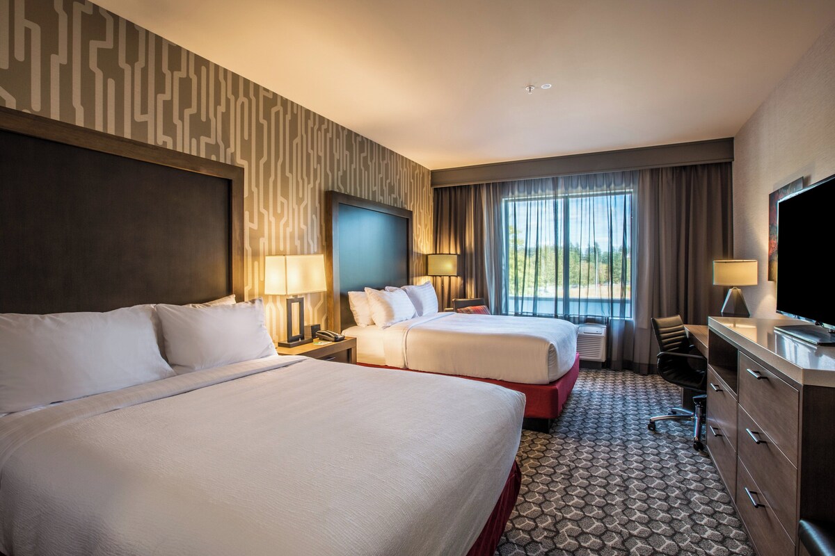 Een hotelkamer met twee bedden, een bureau met een televisie, een stoel en een raam met gordijnen.
