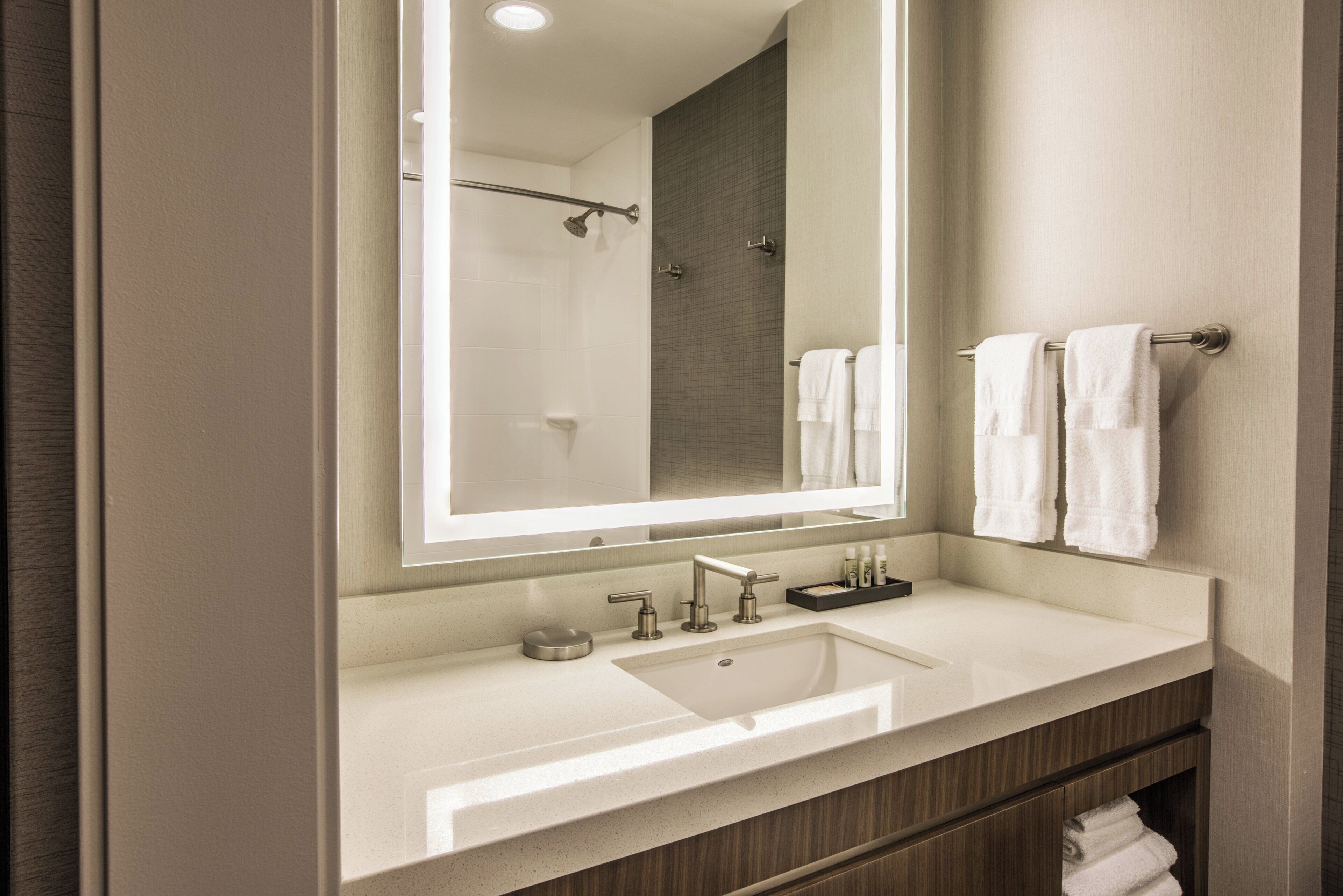 A modern bathroom with a large mirror, a sink, and a shower visible in the reflection.
