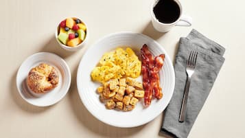 A breakfast plate with scrambled eggs, bacon, and hash browns, a bowl of fruit, and a muffin.