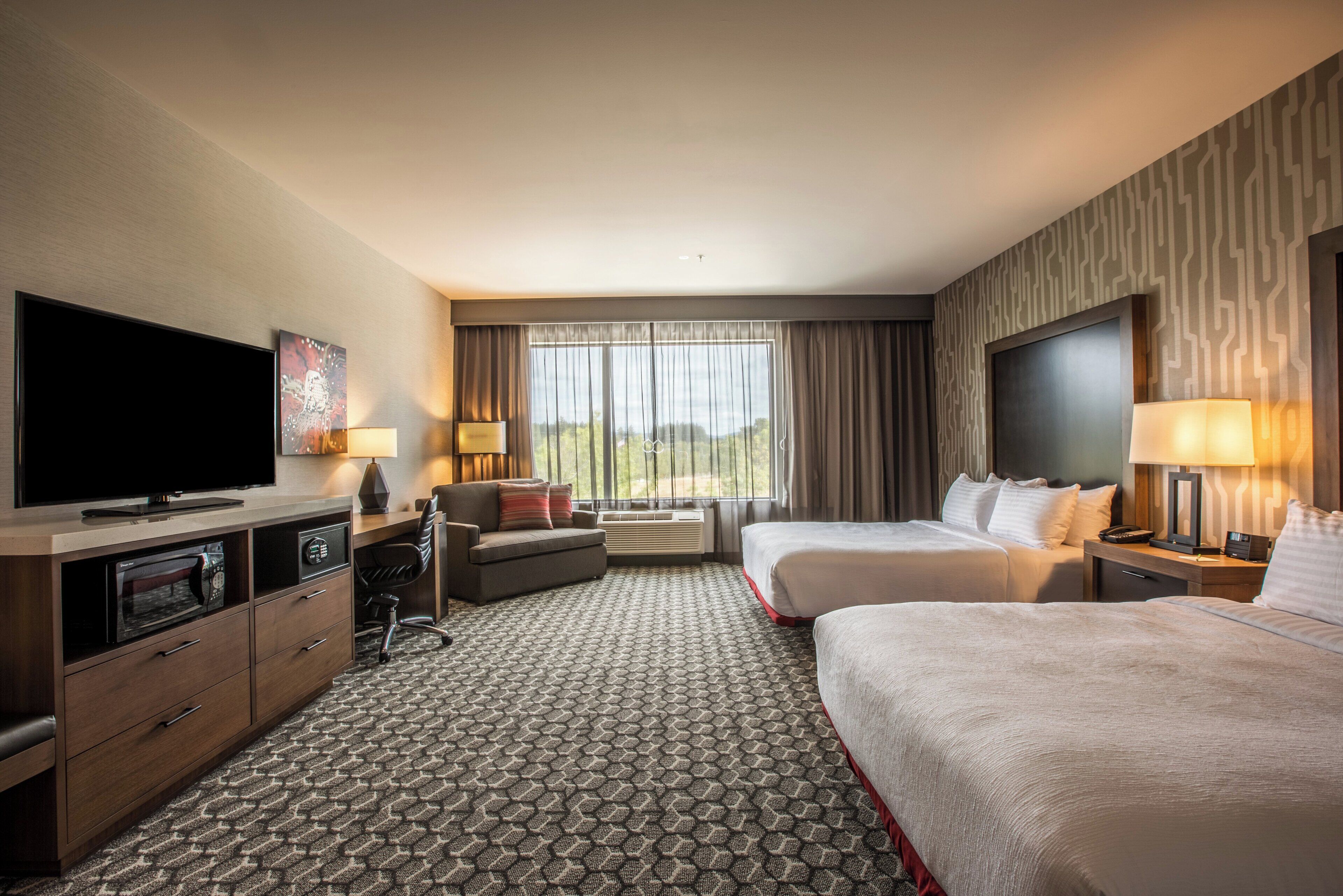 A hotel room with a flat-screen TV, a desk with a computer, two beds, a sofa, and a large window with curtains.