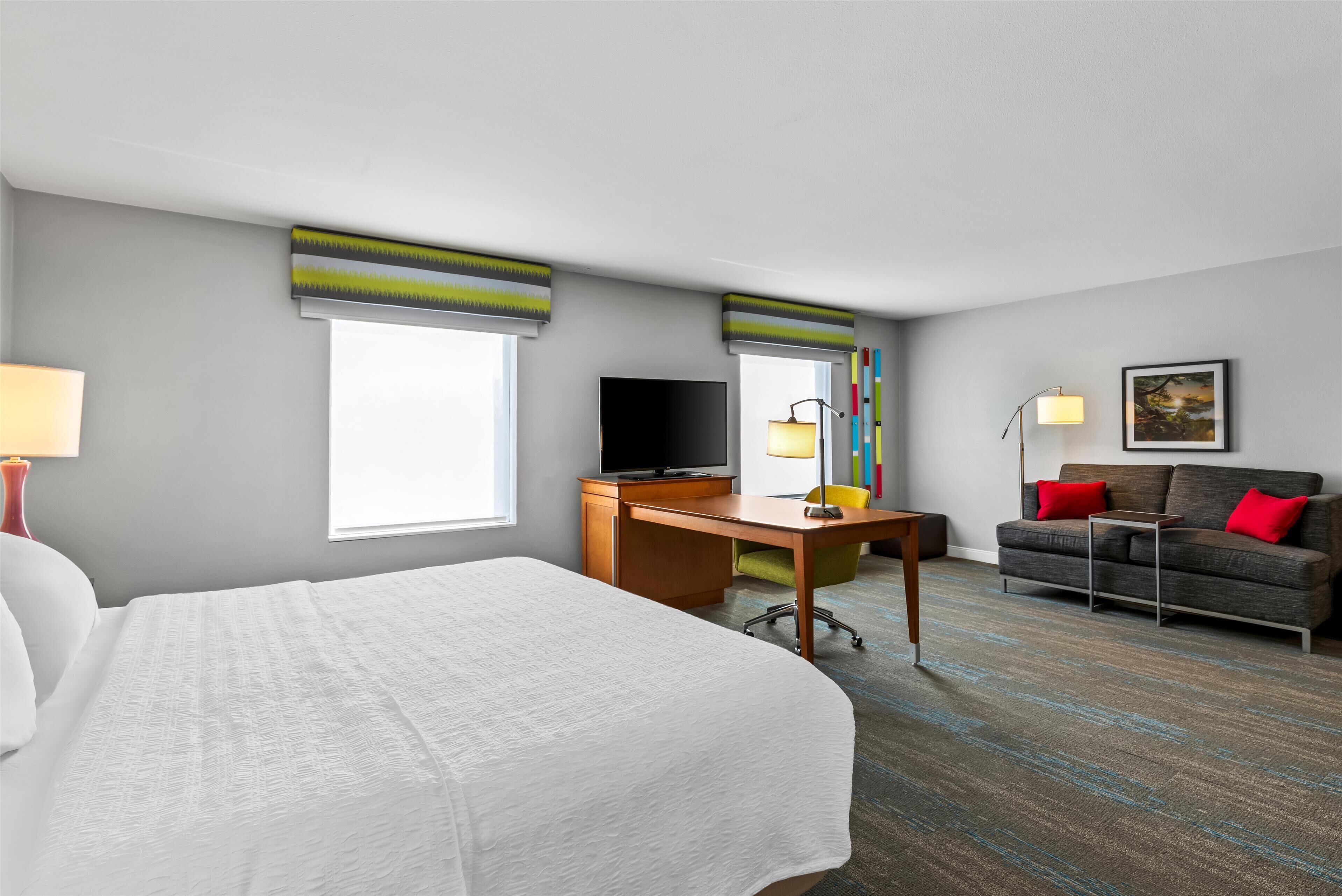 Une chambre d’hôtel équipée d’un lit, d’un bureau, d’une télévision, d’un canapé et d’une fenêtre avec des stores.