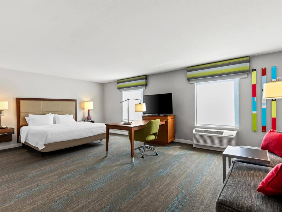 Habitación de hotel con cama, escritorio, silla, televisor y ventana con persianas.