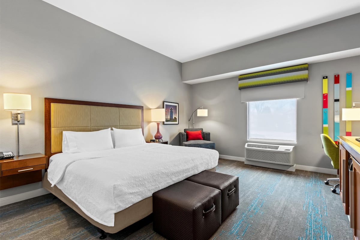 A hotel room with a large bed, bedside tables, a desk, a chair, a window, and an air conditioning unit.