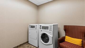 A laundry room with two front-loading washing machines, a brown wicker chair with a yellow cushion, and a tiled floor.