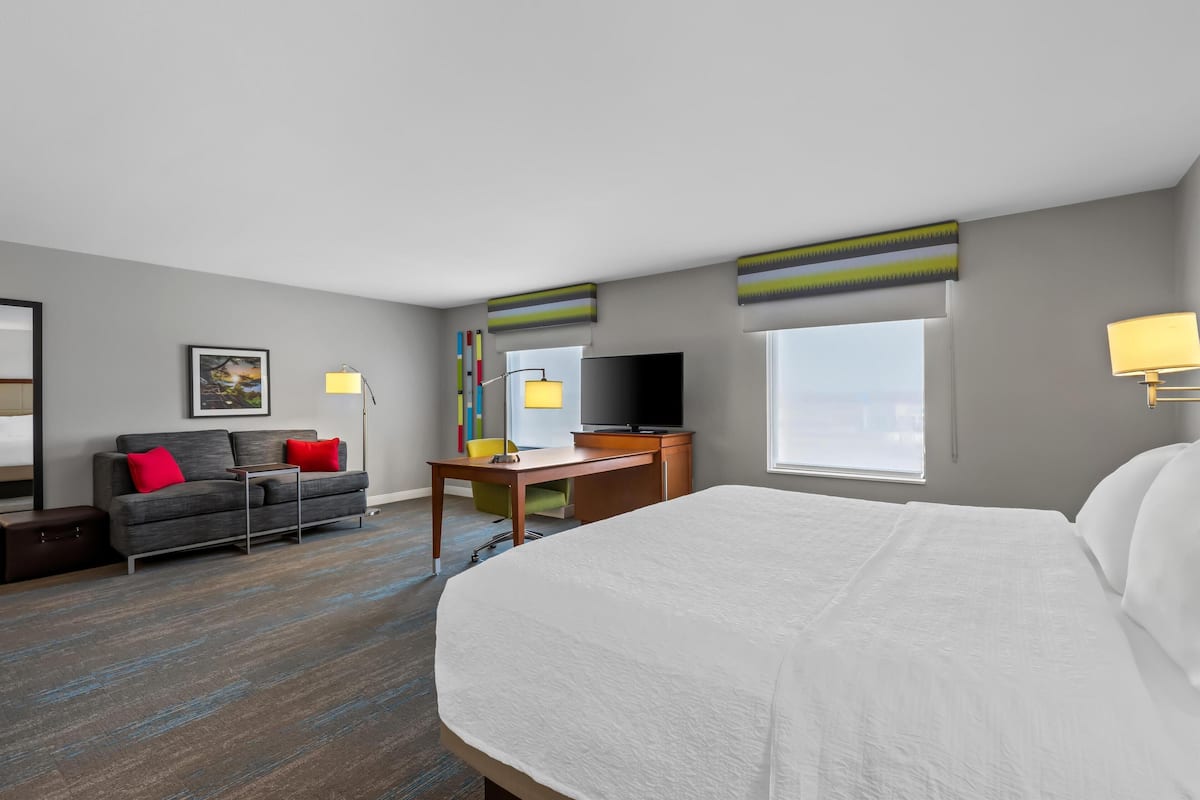 A hotel room with a large bed, a desk with a chair, a sofa with red cushions, a television, and a window with a view.