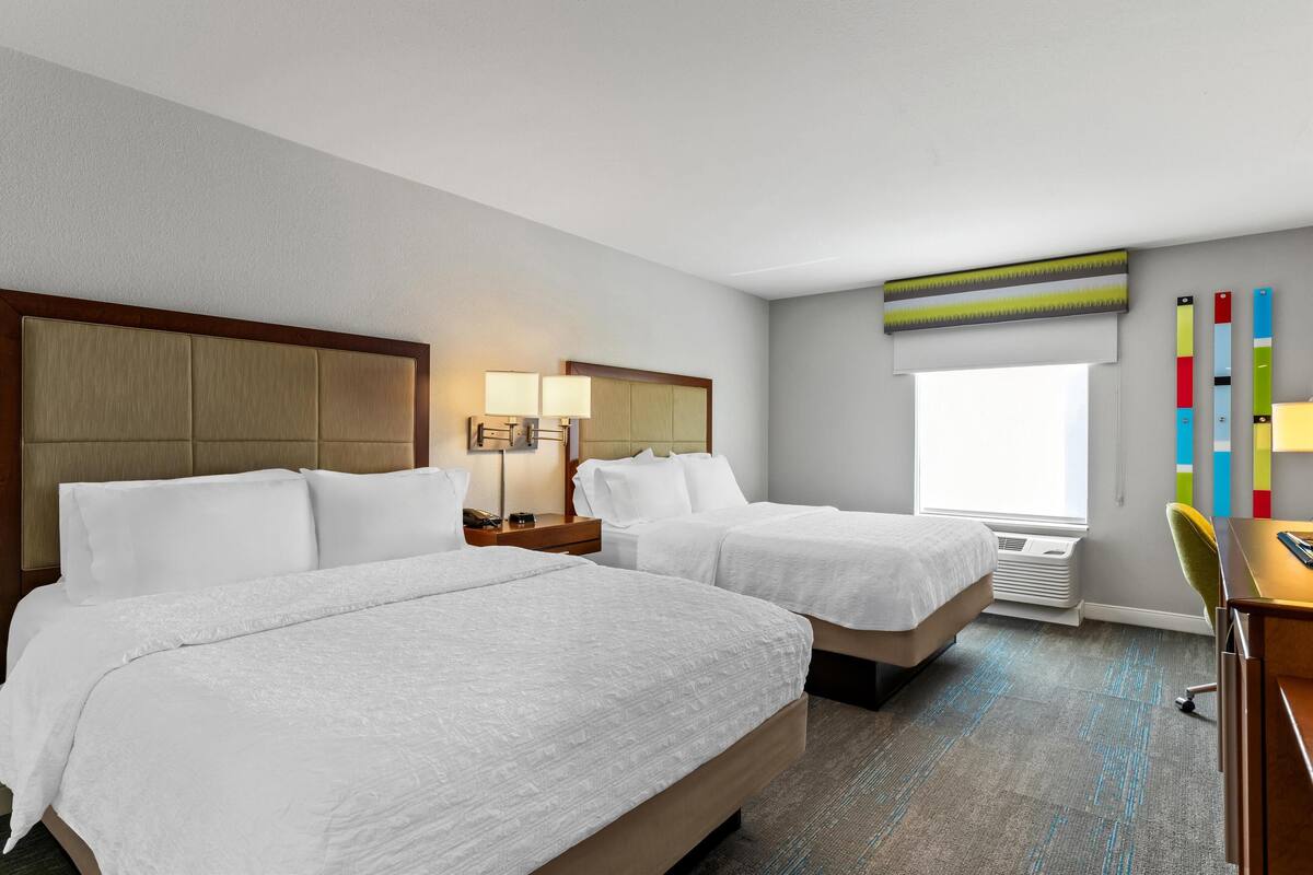 A hotel room with two beds, a desk, a chair, a lamp, and a window with a view.
