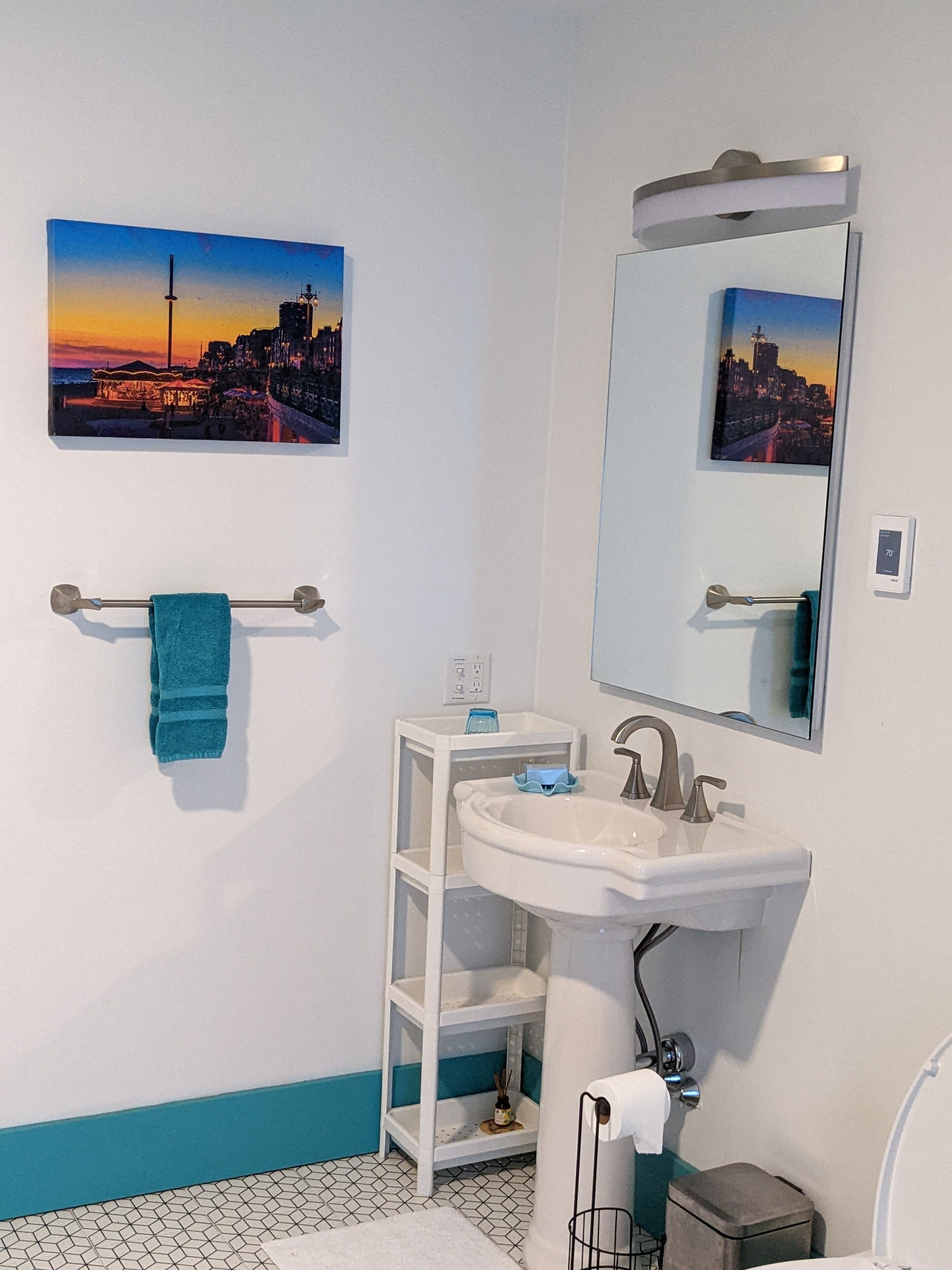 A bathroom with a white sink, a mirror, a towel rack with a blue towel, a small shelf, and a picture of a sunset cityscape on the wall.