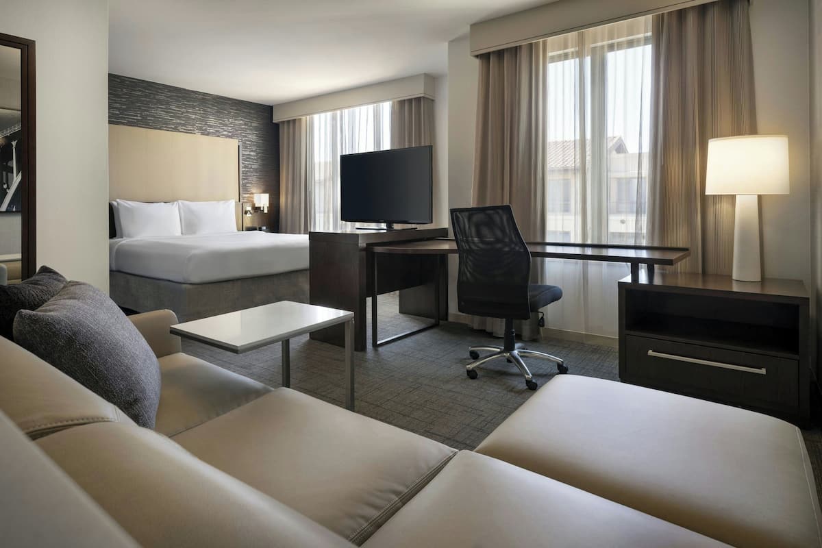 A hotel room with a bed, a desk with a TV, a chair, a small table, and a lamp.